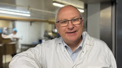 John Foster, Managing Director of Foster's Bakery standing next to a bread oven in his bakery in Barnsley. His wearing a white overall.