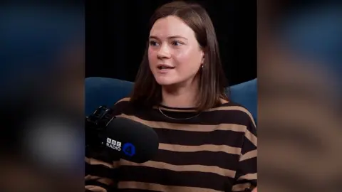 A woman in a brown striped jumper sits on a blue sofa in front of a microphone