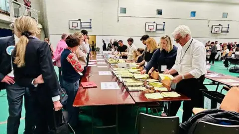 Wide view inside a large sports hall where rows of tables are set up and election staff sort yellow ballot papers, with basketball hoops visible on the walls