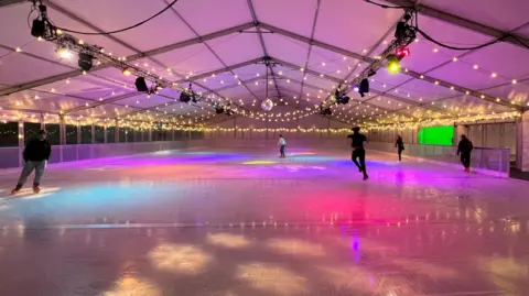 Five people go around an ice rink in Milton Keynes. It is inside a large white marquee and with multi-coloured disco lights shining onto the ice. A large disco ball is hanging above the centre of the rink.