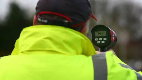 Getty Images An over-the-shoulder photo of someone wearing a cap and high-vis jacket while holding a speed gun. The gun's display reads "34.1"