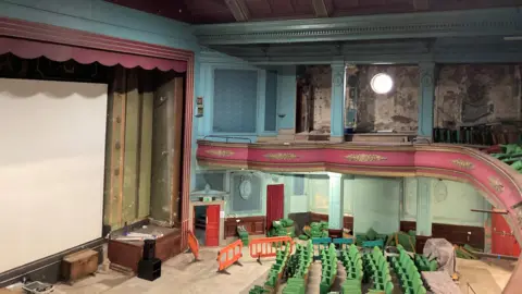 Theatres Trust Abbeydale Picture House auditorium with a large white screen and blue painted walls with pink trim. There is a balcony. There are green seats and orange construction barriers as work progresses.