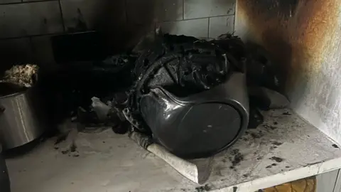 A fire-damaged air fryer on the corner of a kitchen counter showing fire and smoke damaged walls and a melted appliance