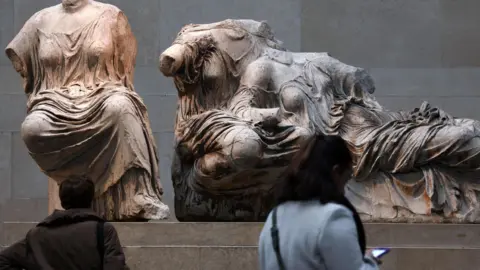 EPA Part of the Elgin marbles moved while two women were looking at them. One person is wearing a brown cropped shirt and the other is wearing a light blue jacket, looking at his phone.