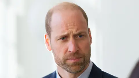 PA Media Prince William looks into the distance at an event while wearing a dark blazer and a light blue shirt. He has a short beard covering his lower face. He is stood in front of a white wall.