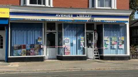 The photo shows a building with a sign saying 'Avenues Library' in white lettering on a blue background above three large windows, which all have vertical blinds drawn behind them. Various posters are stuck on the windows. Between the windows are two white doors with red and white striped tape across their entrances. A man is standing on a step ladder in one of the entrances.