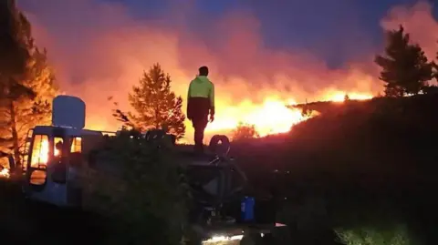 A man standing on a hill with flames in front of him. The sky is lit up orange