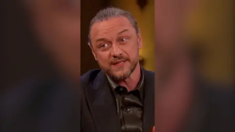 James McAvoy has greying hair tied back, and a beard and moustache. He is wearing a shiny black shirt and a black jacket.