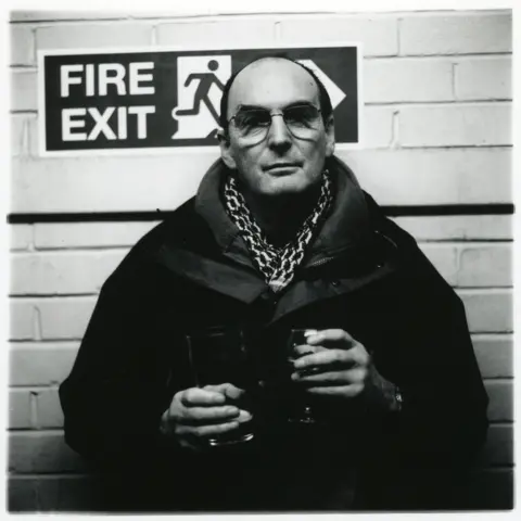 Jason Scott Tilley/Lower Block A black and white portrait of Jason Scott Tilley's father Roy. He is wearing glasses and a scarf and holding a drink in each hand. He is standing in front of a fire exit sign. 