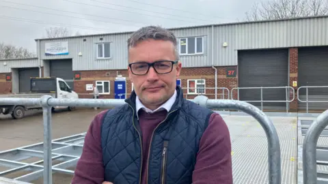 Chris Escott smiling at the camera outside Berry & Escott Engineering. Wearing dark rimmed glasses and a blue body warmer