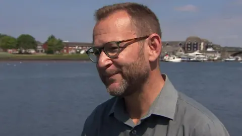 Dr Dave Rumble stands looking slightly off camera as he gives a TV interview. Behind him is the River Itchen, a large expanse of blue water with some greenery and a boathouse in the background and a few white yachts docked on the water. Dr Rumble has short light brown hair and a short beard, with tortoiseshell round thick framed glasses and a grey collared shirt.