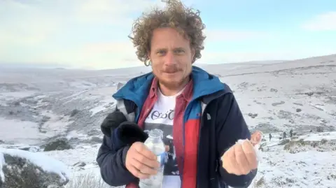 The Corser family Ben Corser is standing in front of a snowy mountainous backdrop. He is smiling and has light brown curly hair. He is holding a bottle of water in one hand and a has a handful of snow in the other which he is stretching towards the camera.
