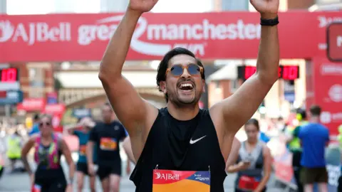 A man smiling with his arms in the air celebrating. He is wearing sun glasses and a black vest top.