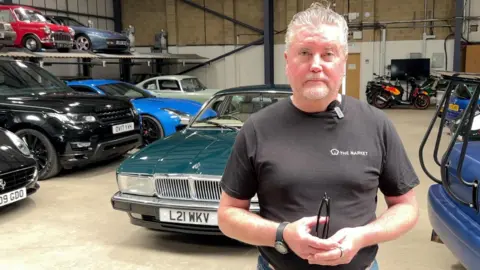 Mark Livesey has short white hair and a black t-shirt on. He is standing in front of dozens of cars in a warehouse.