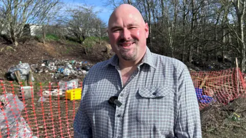 Kate Bradbrook/BBC Richard Johnstone is looking at the camera and standing outside on a sunny day. He is balding and has facial hair. He is wearing a blue checked shirt and standing in front of a site covered in litter.