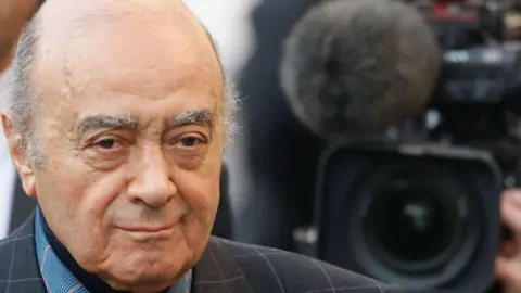 Getty Images Mohamed Al Fayed with mild smirk. He is looking straight on. His eyebrows are fairly bushy and he is balding on the top of his head. He is wearing a black suit jacket with a thin checked pattern on. The photo was taken in 2008.