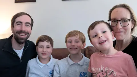 Calum Dad Calum, his sons Archie, Andrew, Roddy and mum Katie sit next to each other on a sofa and smile at the camera. Archie and Andrew are in their school uniforms and Roddy is wearing a top with monkeys on it. 