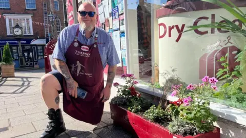 Ellen Knight/BBC Kenny Bould pictured kneeling down next to his planter - a long, wooden structure painted dark red with pink flowers planted in it. Bould is wearing a blue check shirt, dark shorts, and a dark red Timpson's-branded apron over the top. There is strong sunshine, and the shop itself can be seen behind the planter. 
