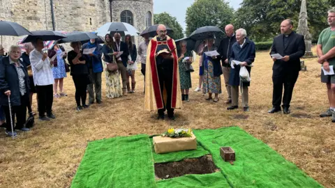 Hannah Gray/BBC A reverend and a group of other people standing in an area during a ceremony. There is a parcel, wrapped in brown paper and flowers attached to it. It is preparing to be buried. 