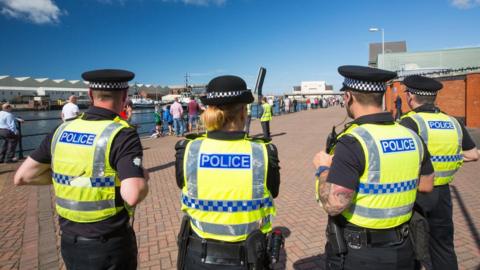 Four police officers on patrol in Barrow, with their backs to the camera near a riverside location with people lined up long the water's edge