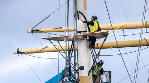 Martin Shields Two men in high vis yellow jackets, working on the mast of a large boat. They are helping to remove the masts for conservation work.