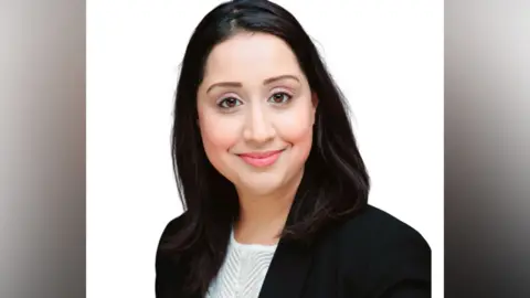 Maryam Yaqub A woman with long dark hair smiles at the camera. She wears a black suit jacket with a white top underneath. 