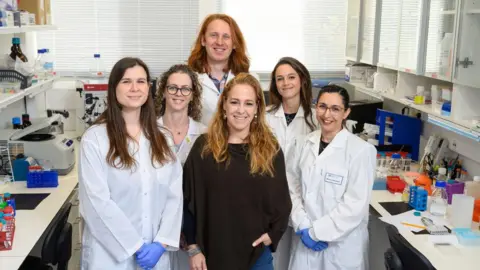 El Grupo de Científicos del Instituto Weizmann de seis científicos, cinco mujeres y un hombre, tomando una foto grupal, la mayoría usa abrigos de laboratorio blanco en un entorno de laboratorio.