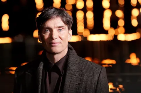 Actor Cillian Murphy stares into camera with a gentle smile on his face. He's wearing a brown coat and burgundy silk shirt. 