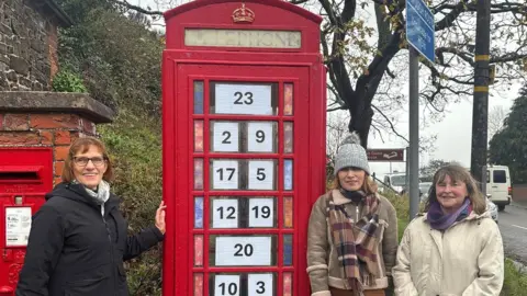 Three women standing next to a red phone box. In each window is a piece of paper with a black number on it. Next to the phone box is a red post box - they both sit next to a pavement and road