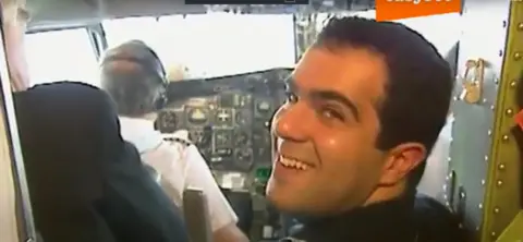 easyGroup Stelios stands in the doorway of the cockpit and smiles back at the camera. We can see the back of a pilot or co-pilot sitting on his chair and wearing a white short sleeved shirt and headphones.