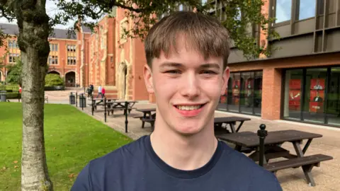 Jake is standing in front of a tree and benches. He has floppy brown hair and is wearing a navy blue tshirt. He is smiling at the camera.