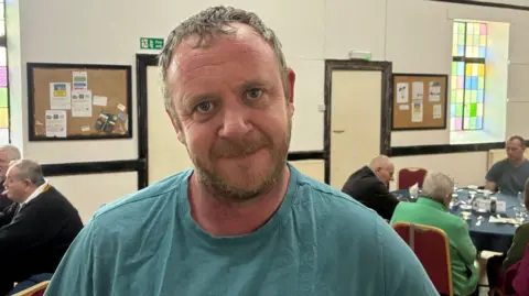 The image shows a man with a beard, wearing a greenish blue T-shirt, looking at the camera 