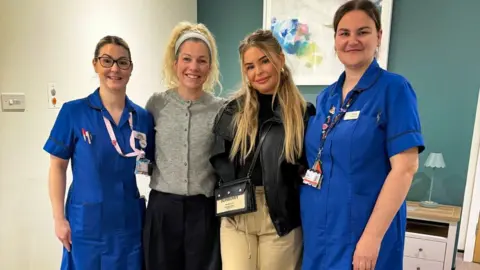 WISHH Fundraisers Juliet O'Sullivan and Sasha Larsen pictured with two members of the midwifery bereavement team, who are wearing blue hospital nurse's uniforms. They are standing inside a refurbished unit in front of a picture on the wall.
