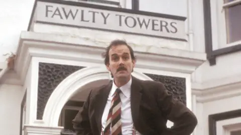 A man with a moustache wearing a suit in front of a hotel saying Fawlty Towers