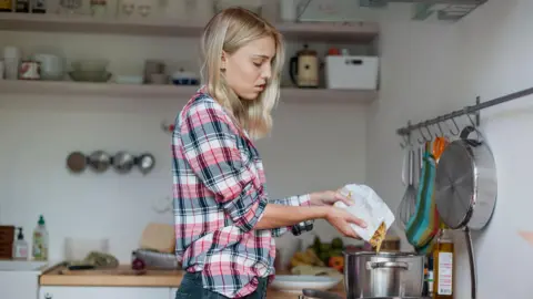 A young woman with shoulder length blonde hair wearing a navy red and white check shirt pouring pasta into a silver pan on a stove in a basic looking kitchen