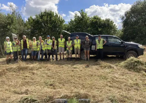 A group of volunteers wearing hi-vis vests at a previous event hosted by Torridge District Council. The group are standing in a field beside a vehicle and there are trees behind them.