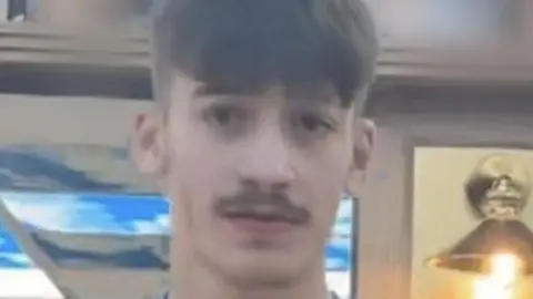 Taylor Charlton is wearing a blue shirt with a white Nike logo on it. He has a short moustache and short hair.