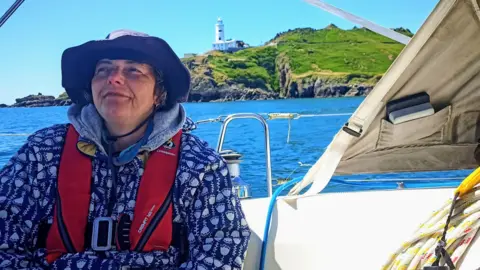 Antje Ronneberger The picture shows a person sitting on a sailing boat on a bright, clear day at sea. The person is in the foreground, wrapped up in a patterned jacket and wearing a wide-brimmed hat, with a red life jacket fastened across the chest. The boat’s cockpit surrounds them, with ropes neatly coiled to one side and parts of the canvas canopy and rigging framing the shot.