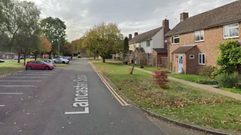 Google Maps Lancaster Square in Lyneham. There are rows of parking spaces to the left, with residential properties to the right and a grassy verge.