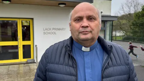 Canon Alan Irwin is standing in front of the entrance to  Lack Primary School. He is a pastor, so is wearing a collar. His clothes are navy. Children are playing behind him.