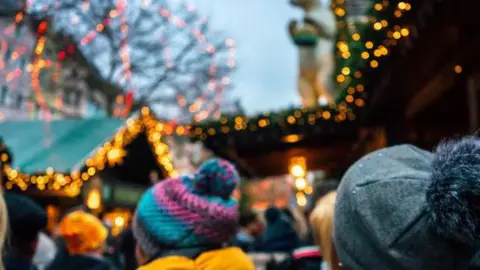 The back of people's head in bobble hats. They are at a Christmas fair. There are blurred fairy lights on market stalls and in a tree with no leaves.
