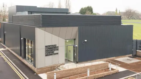 The exterior of the diagnostics unit, it has grey cladding, there is a churchyard in the background.