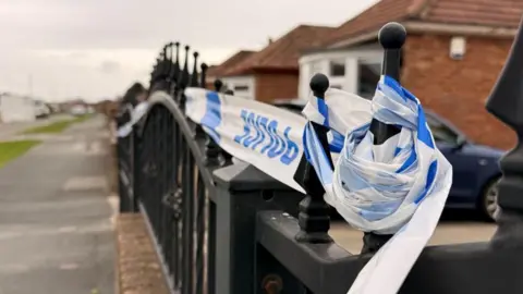 The image shows a residential street lined with single‑storey houses. The focus is on the front boundary fence of one property, where a section of blue‑and‑white tape is tied around the metal railings.