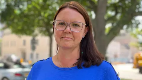 Emma is wearing glasses and a blue top standing outdoors on a tree-lined street, with parked cars and buildings blurred in the background.