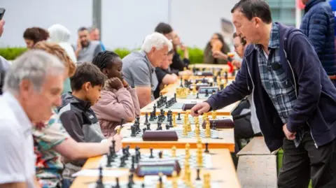 Chess in Schools and Communities A row of tables with chess boards and on with a miox of children and adults playing the game