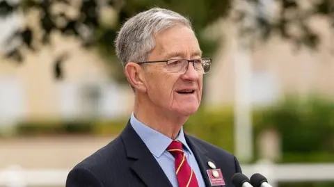 Richard Palusinksi stands in front of two microphones as he delivers a speech. He has short grey hair and is wearing glasses, a blue shirt, red tie and navy-blue jacket with medals displayed on his right breast.