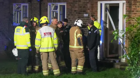 Eddie Mitchell A group of fire fighters and police officers gather outside the door of a block of flats that has caught fire in Steyning. The front door is open but sealed off by police tape.