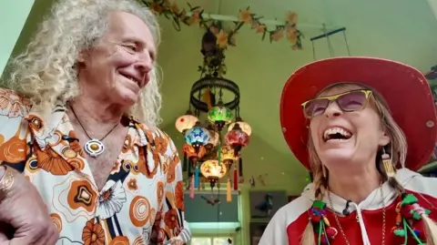 A tall man with curly blond hair looks down at a laughing woman wearing a cowboy hat and glasses and her hair in pigtails
