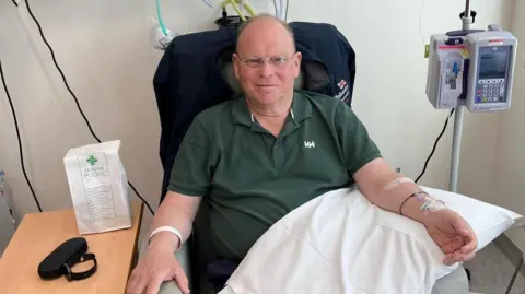 Andy Gissing sits in a hospital chair with a tube in his arm, which rests on a pillow on his lap. He is mostly bald and wears glasses and a green polo shirt.