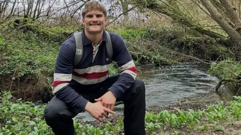 James Hirst Man crouched in hiking gear on a grassy path. He has dark trousers on, light brown hair, and is carrying a rucksack